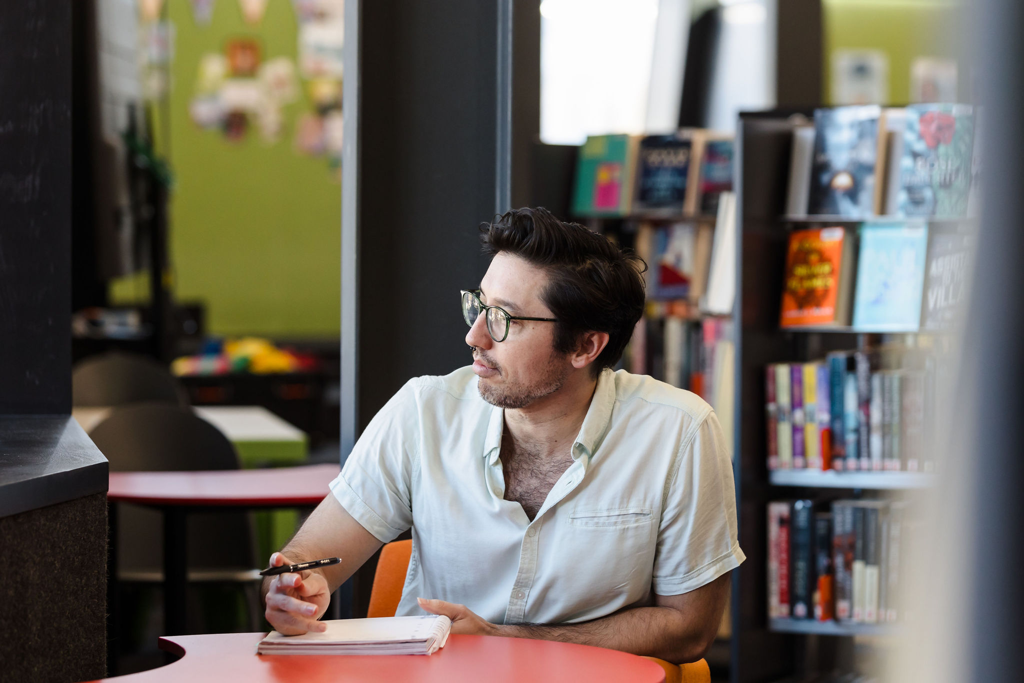 A person sits at a red table in a library, holding a pen and looking thoughtfully to the side. Bookshelves filled with colorful titles are visible in the background, creating a calm and focused atmosphere.